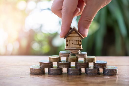 Haus in 2 Fingern gehalten und auf Pyramide aus Geld gesetzt